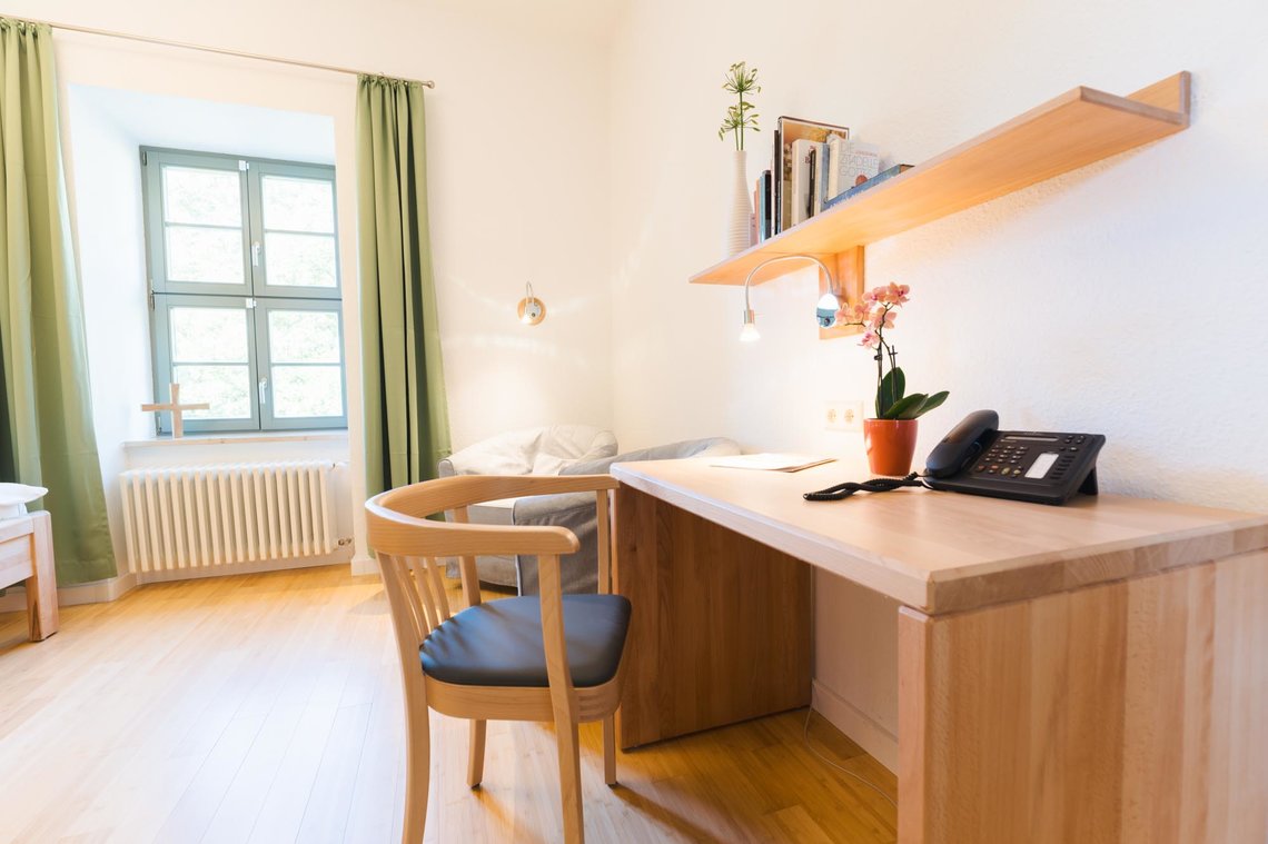 Ein helles weißes Zimmer mit einem Fenster mit grünen Vorhängen. Im Vordergrund an der rechten Wand steht ein Schreibtisch mit einem Stuhl. Auf dem Schreibtisch steht ein Telefon und eine Blume. Über dem Schreibtisch ist ein Bücherregal unter dem eine Lampe hängt. Hinter dem Schreibtisch stehen zwei graue Sessel.