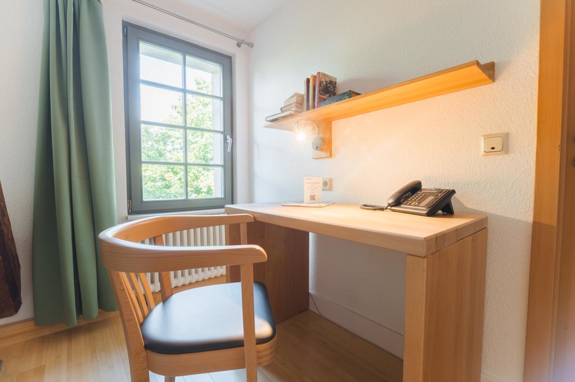 Ein Stuhl steht vor einem Schreibtisch, auf dem ein Telefon steht. Darüber hängt ein Bücherregal