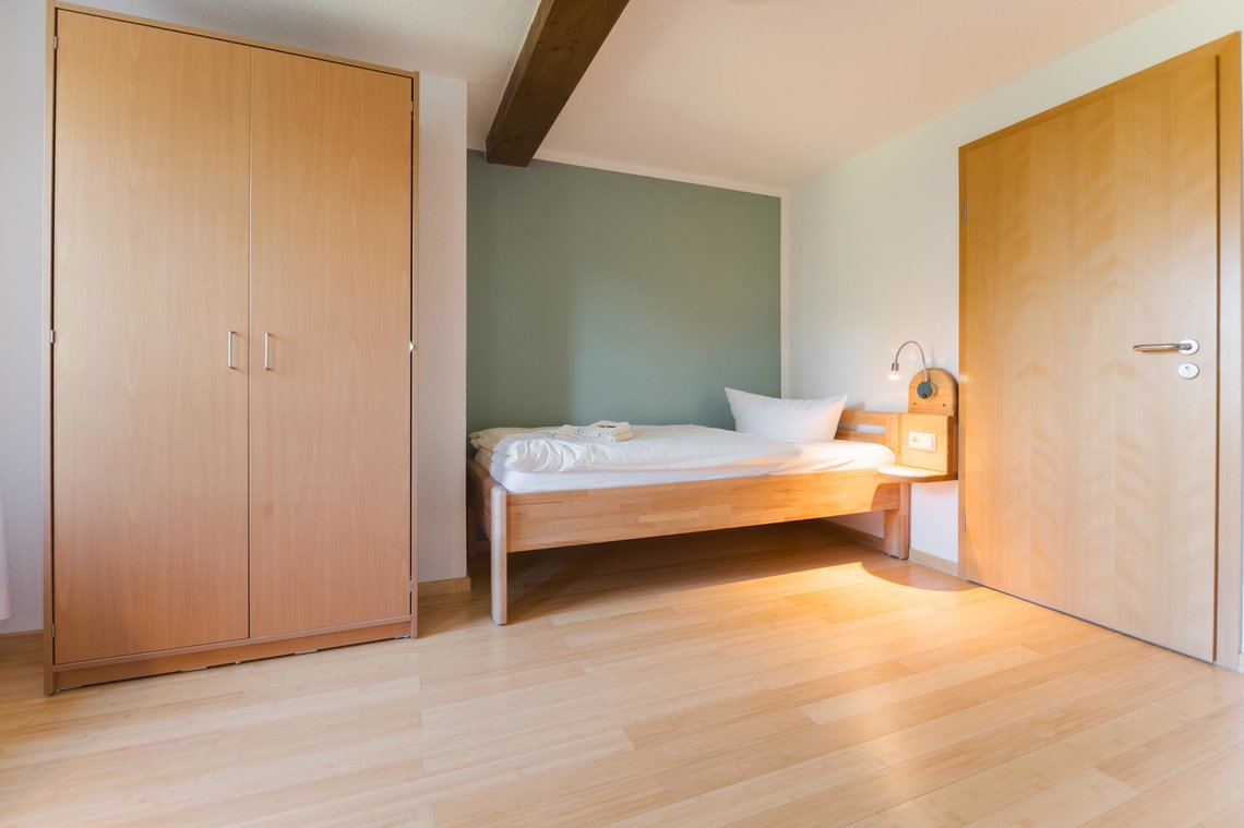 Ein Bett steht neben einer grünlichen Wand neben einer Tür. Auf dem Bett liegen zwei Handtücher und neben dem Bett ist eine Lampe mit einer Ablage. Links neben dem Bett steht ein Schrank mit zwei Türen.