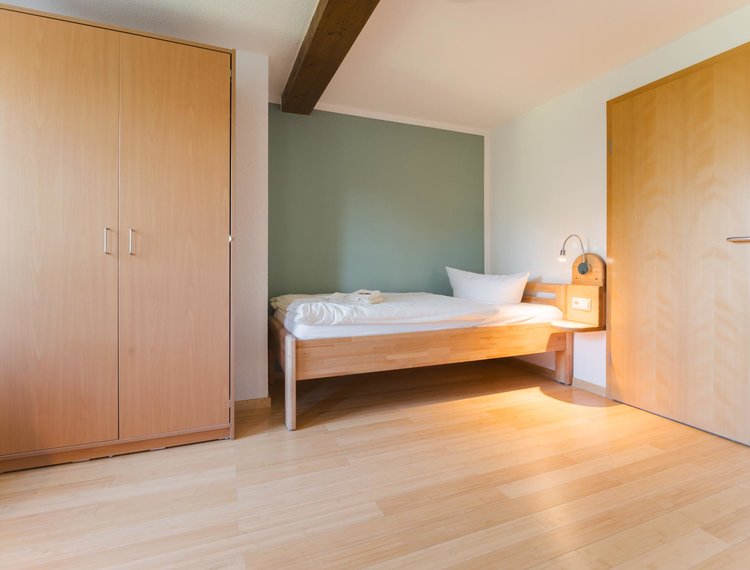 Ein Bett steht neben einer grünlichen Wand neben einer Tür. Auf dem Bett liegen zwei Handtücher und neben dem Bett ist eine Lampe mit einer Ablage. Links neben dem Bett steht ein Schrank mit zwei Türen.