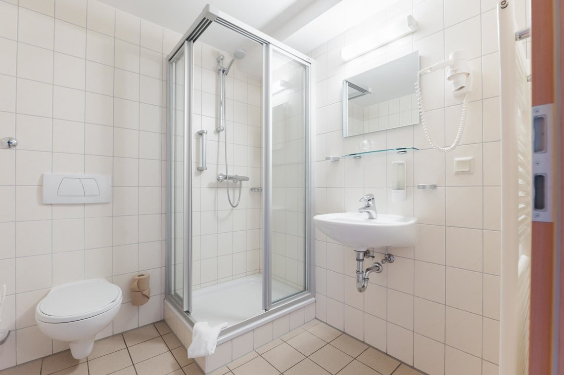 Ein Badezimmer mit einer Dusche in der Ecke. Am Rand der Dusche liegt ein Handtuch. Links neben der Dusche befindet sich eine Toilette und rechts neben der Dusche ist ein Waschbecken mit einem Spiegel. Über dem Spiegel ist eine Lampe, rechts daneben ist ein Fön in einer Halterung und unter dem Spiegel ist eine Glasablage.