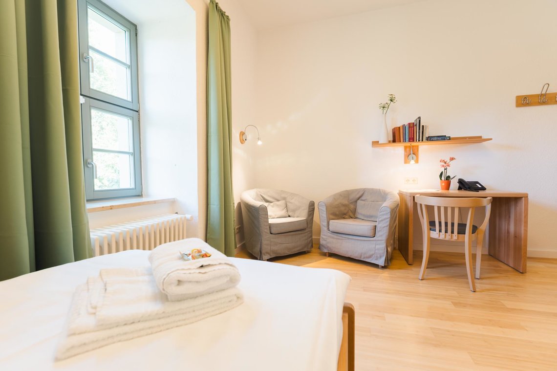 Ein helles weißes Zimmer mit einem Fenster mit grünen Vorhängen. Im Vordergrund steht ein Bett mit 2 Handtüchern und Gummibärchen. An der Wand steht ein Schreibtisch mit einem Stuhl. Auf dem Schreibtisch steht ein Telefon und eine Blume. Über dem Schreibtisch ist ein Bücherregal unter dem eine Lampe hängt. Links neben dem Schreibtisch stehen zwei graue Sessel. 