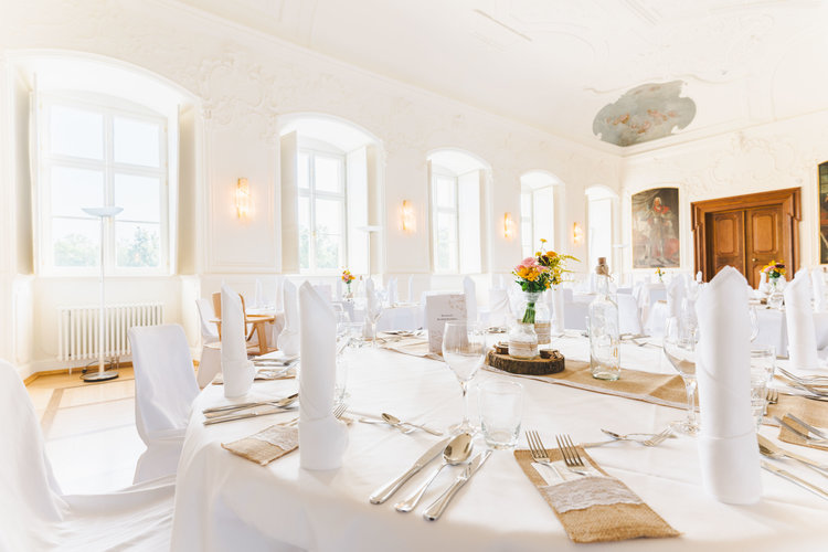 Der große Kaisersaal ist feierlich geschmückt. Die runden Tische sind mit weißen Tischdecken gedeckt und die Stühle in weißen Stoffen gehüllt. Die Tische sind gedeckt mit Besteck, weißen Servietten, einer Blumenvase und der Buffetkarte.