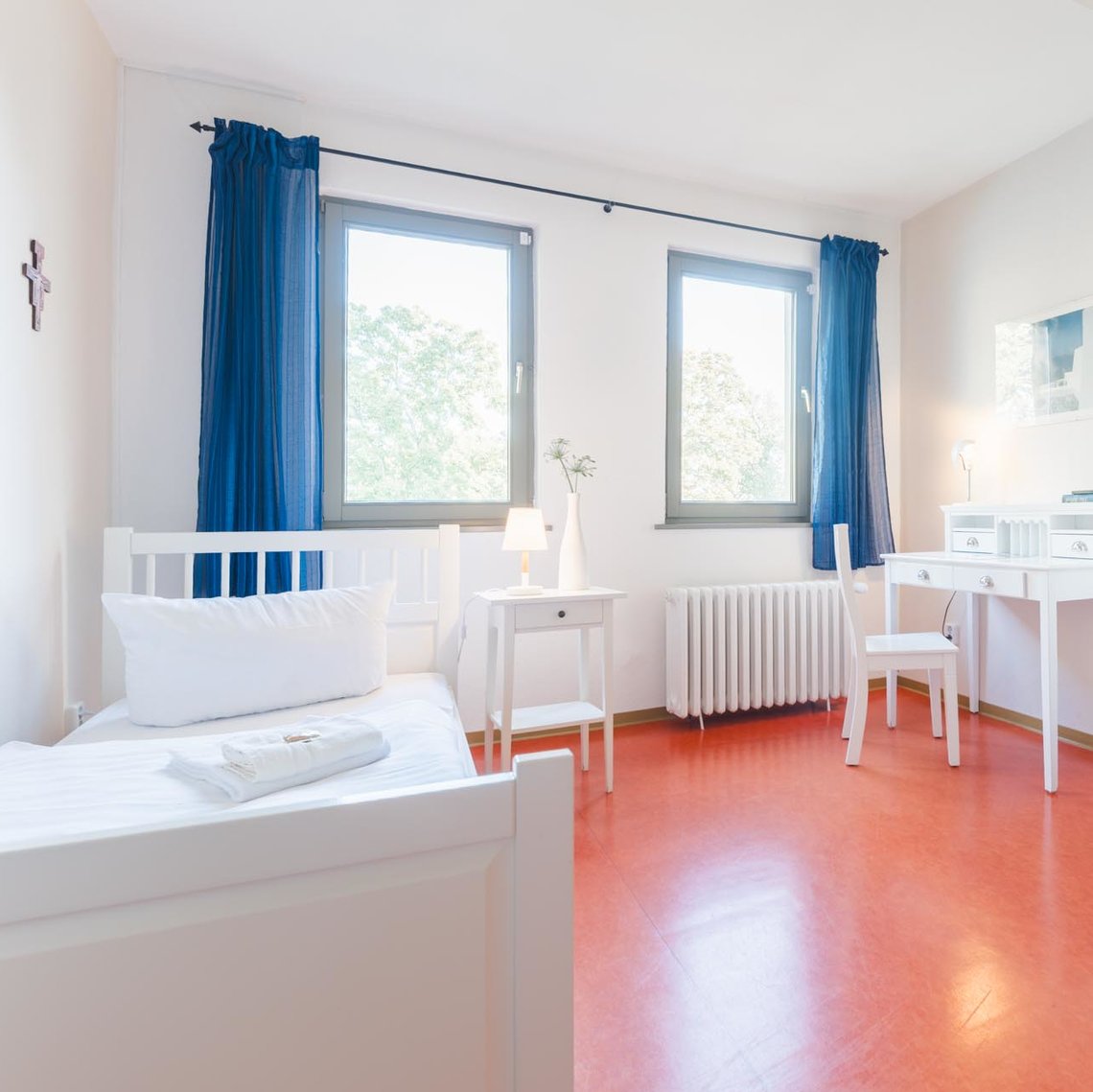 Ein Hotelzimmer mit 2 Fenstern, weißen Wänden, weißen Möbeln, blauen Gardinen und einem roten Boden. Links unter einem Kreuz an der Wand steht ein Bett mit 2 Handtüchern. Daneben steht ein Nachttisch mit einer Lampe und einer Blume. An der Rechten Wand steht ein Schrank und links daneben ein Schreibtisch mit einem Stuhl. Über dem Schreibtisch hängt ein Bild.