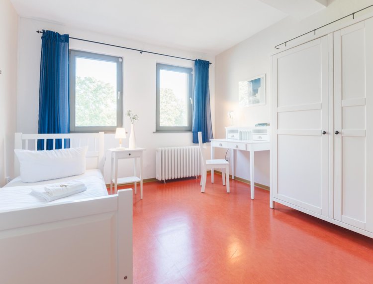 Ein Hotelzimmer mit 2 Fenstern, weißen Wänden, weißen Möbeln, blauen Gardinen und einem roten Boden. Links unter einem Kreuz an der Wand steht ein Bett mit 2 Handtüchern. Daneben steht ein Nachttisch mit einer Lampe und einer Blume. An der Rechten Wand steht ein Schrank und links daneben ein Schreibtisch mit einem Stuhl. Über dem Schreibtisch hängt ein Bild.