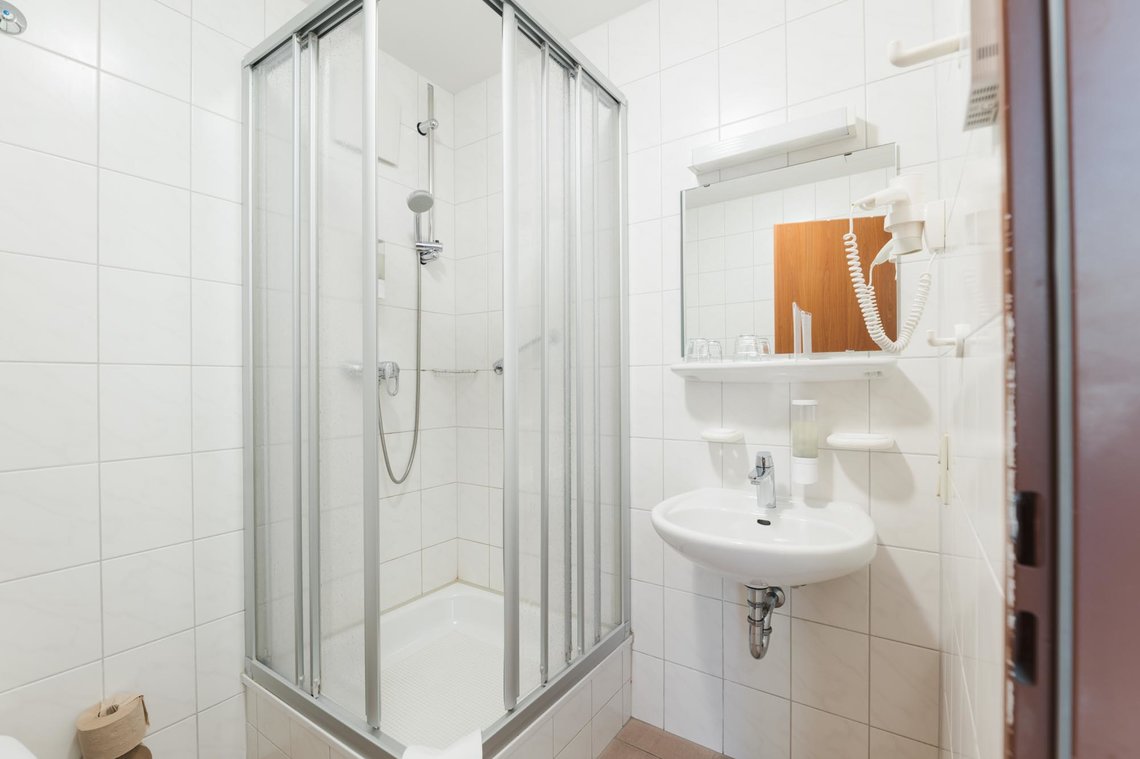 Ein kleines weiß gefliestes Bad mit einer Dusche in der Ecke und einem Waschbecken mit Spiegel, Ablage und Fön.