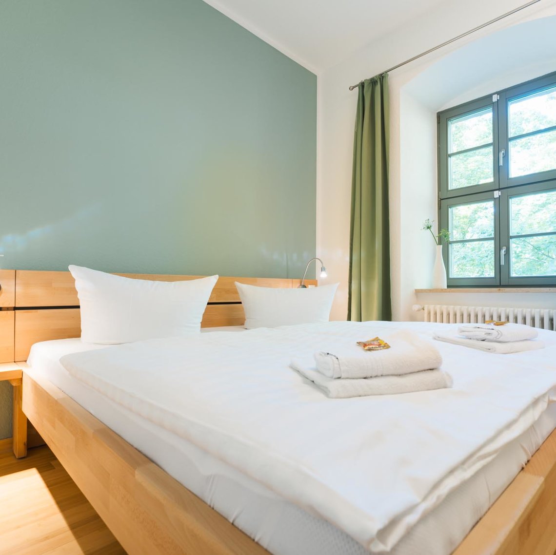 Ein Hotelzimmer mit einer weißen und einer grünen Wand und 2 Fenstern mit grünen Vorhängen. Links vor der grünen Wand steht ein Doppelbett mit zwei Lampen. 