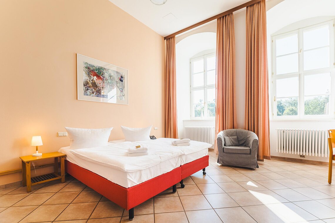 Ein großes Doppelbett und ein Sessel stehen in einem geräumigen Raum mit hohen Fenstern.