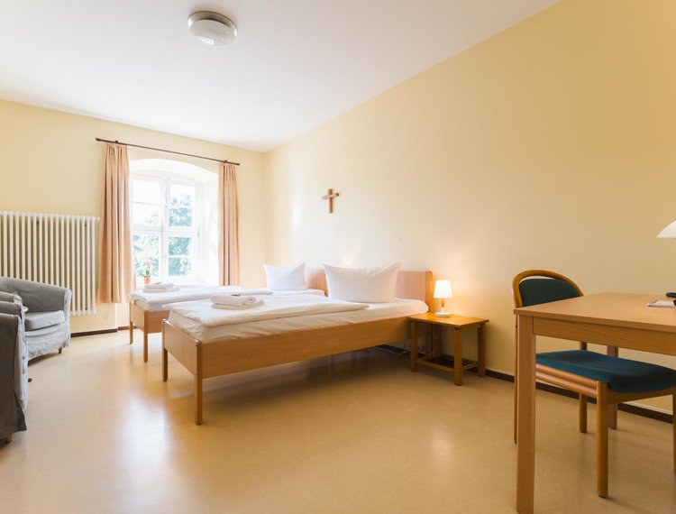 Ein schlichtes, helles Zweibettzimmer. In der Mitte stehen zwei Einzelbetten mit weißer Bettwäsche und einem kleinen Nachttisch mit Lampe dazwischen. Links befinden sich zwei graue Sessel, rechts ein großer Schreibtisch mit Stuhl und einer Tischlampe. Ein großes Fenster mit Vorhängen lässt Tageslicht herein, an der Wand hängt ein Kreuz