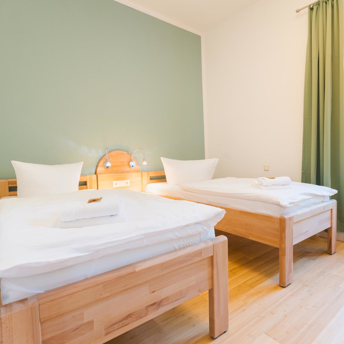 Ein helles weißes Zimmer mit einem Fenster mit grünen Vorhängen. An einer grünen Wand stehen zwei Einzelbetten mit zwei Lampen dazwischen.