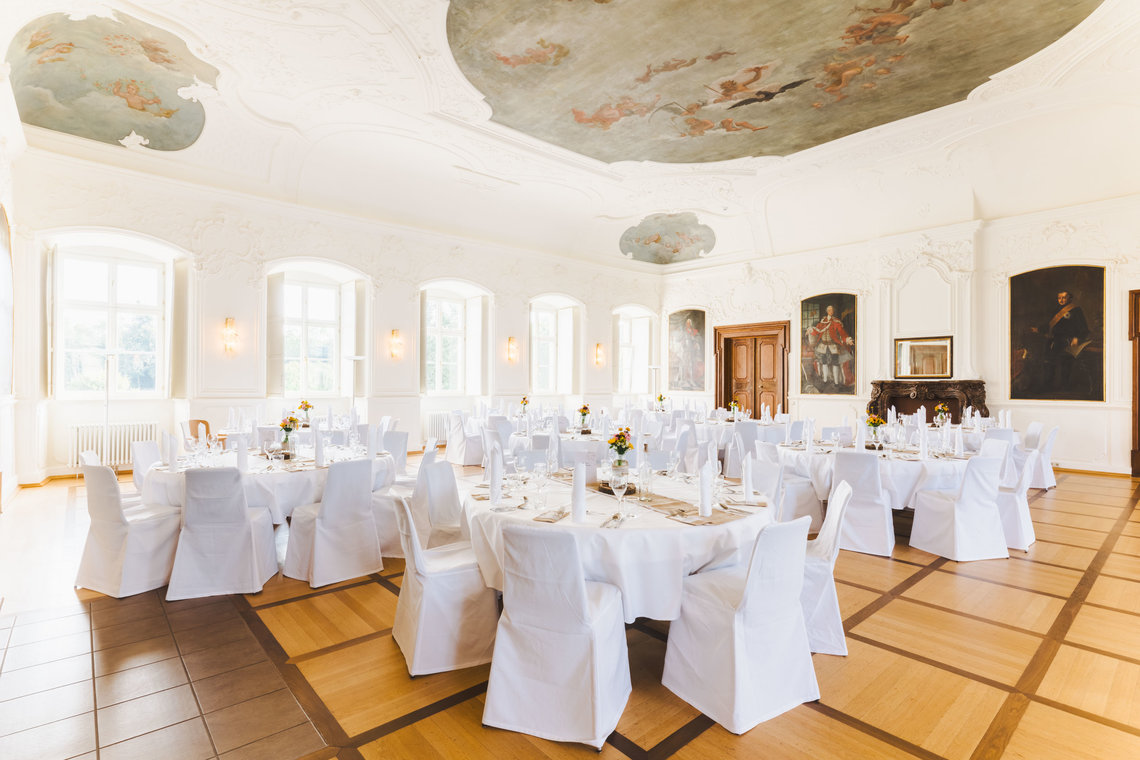 Der große Kaisersaal ist feierlich geschmückt. Die runden Tische sind mit weißen Tischdecken gedeckt und die Stühle in weißen Stoffen gehüllt. Die Tische sind gedeckt mit Besteck, weißen Servietten und einer Blumenvase