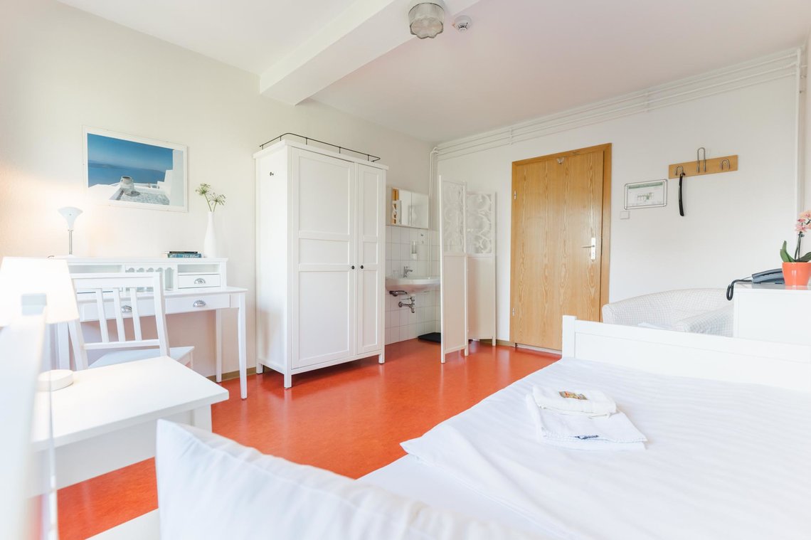 Ein Hotelzimmer mit weißen Wänden, weißen Möbeln und einem roten Boden. Aus der Sicht eines Bettes auf dem 2 Handtücher liegen sieht man von links nach rechts: einen Nachtschrank mit einer Lampe, einen Schreibtisch mit einem Stuhl, einer Lampe, einer Blume, einer Bibel und einem Bild darüber, einen Schrank, daneben ein Waschbecken mit Spiegel hinter einem Sichtschutz, eine Tür, daneben ein Notfallplan und Kleiderhaken, einen Sessel, neben einem kleinem Schrank mit einem Telefon und einer Blume darauf.