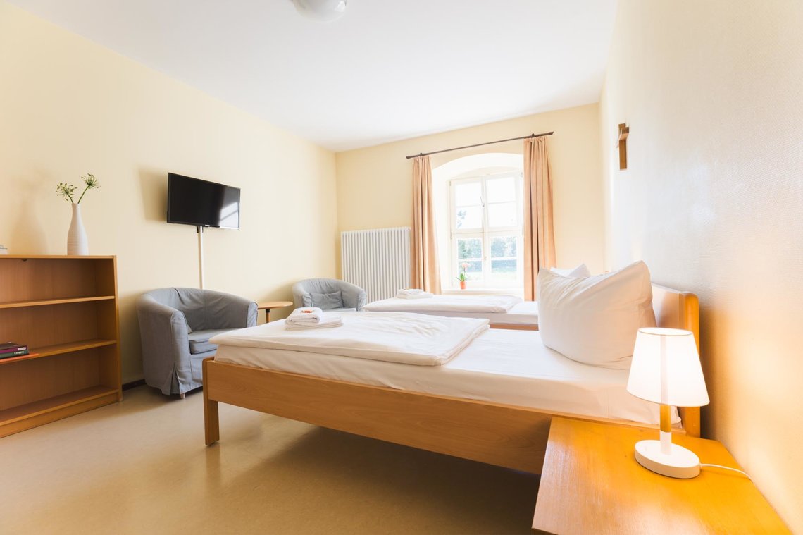 Das Foto zeigt ein schlichtes, helles Zweibettzimmer. An der rechten Wand stehen zwei Einzelbetten mit weißer Bettwäsche und einem kleinen Nachttisch mit Lampe. Links befinden sich zwei graue Sessel, mit neben einem Regal mit einer Blume und einem Beistelltisch. Darüber hängt ein Fernseher an der Wand. Ein großes Fenster mit Vorhängen lässt Tageslicht herein, an der Wand hängt ein Kreuz.