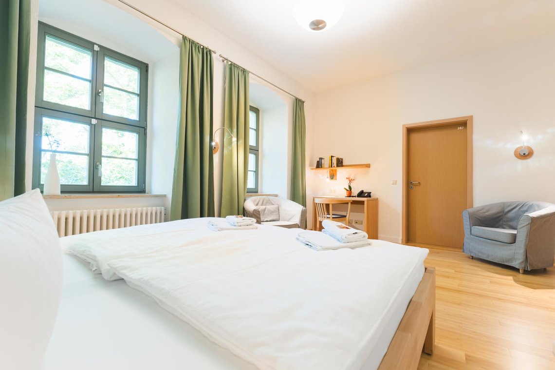Ein Hotelzimmer mit 2 Fenstern mit grünen Vorhängen. Links steht ein Doppelbett mit zwei Lampen. Ein grauer Sessel steht vor dem Fenster. In einem Fenster steht eine Blume. Neben dem Sessel steht ein Schreibtisch mit einem Stuhl. Auf dem Schreibtisch steht eine Blume und ein Telefon. Darüber hängt ein Bücherregal mit einer Lampe. Rechts daneben befindet sich eine Tür und ein weiterer grauer Sessel.