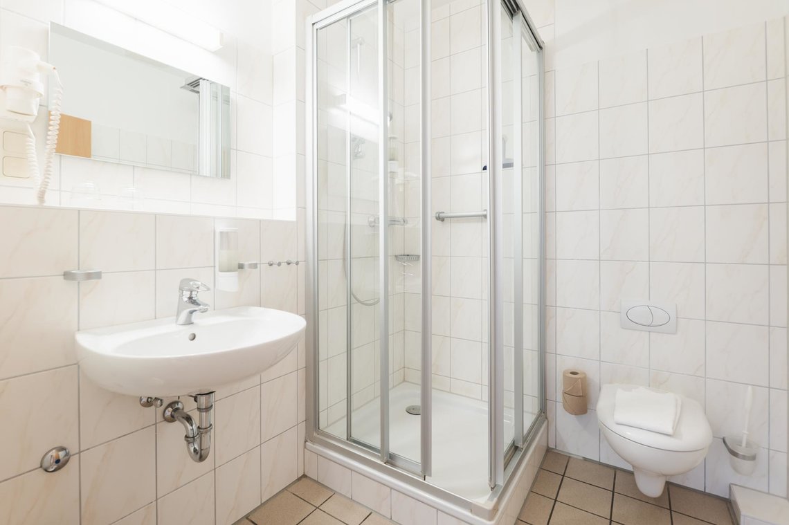 Ein kleines weiß gefliestes Bad mit einer Dusche in der Ecke und einem Waschbecken mit Spiegel, Ablage und Fön. Rechts neben der Dusche ist eine Toilette.