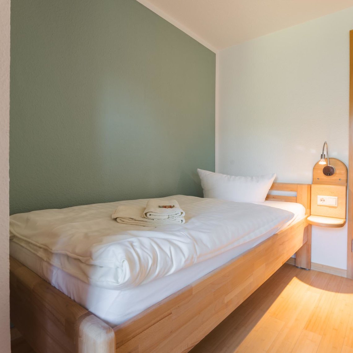 Ein Bett steht neben einer grünlichen Wand neben einer Tür. Auf dem Bett liegen zwei Handtücher und neben dem Bett ist eine Lampe mit einer Ablage.