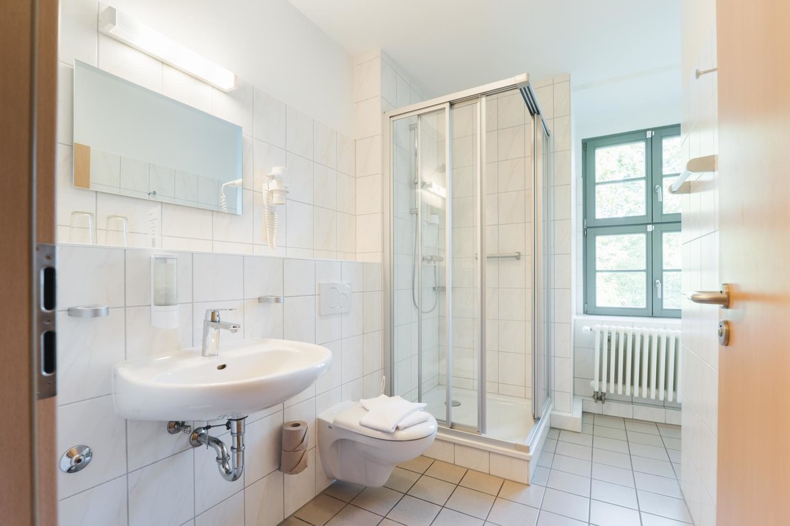 Ein weiß gefliestes Badezimmer mit einem Fenster, einer quadratischen Dusche in der Ecke, einer Toilette mit 2 Handtüchern darauf und einem Waschbecken mit einem Spiegel. Neben dem Spiegel hängt  ein Fön in einer Halterung.